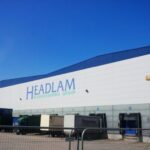 Exterior of a Headlam Floorcovering Group distribution warehouse with loading bays and delivery trucks under a clear blue sky.