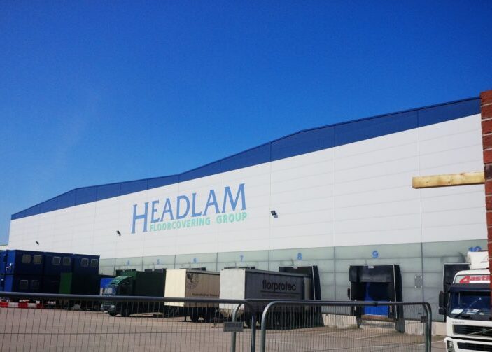 Exterior of a Headlam Floorcovering Group distribution warehouse with loading bays and delivery trucks under a clear blue sky.