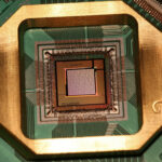 Macro photograph of a D-Wave quantum processor chip, labeled 'D:Wave' and 'Washington', mounted in a gold sample holder on a green circuit board