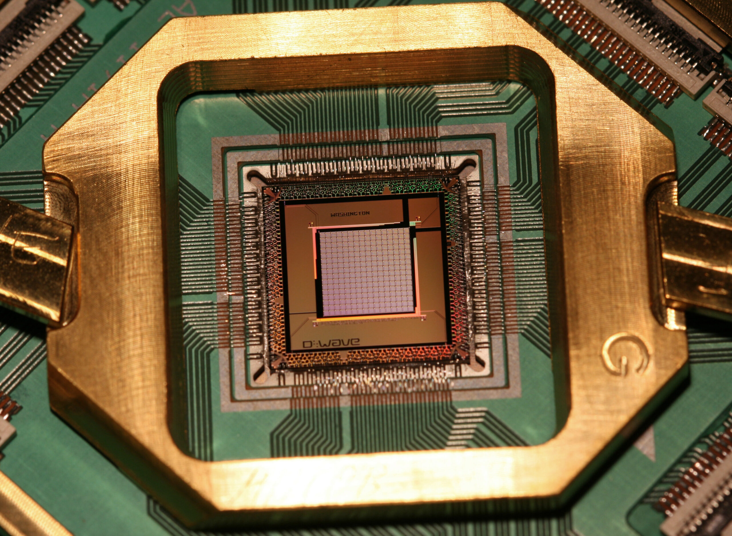 Macro photograph of a D-Wave quantum processor chip, labeled 'D:Wave' and 'Washington', mounted in a gold sample holder on a green circuit board