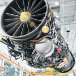 Close-up view of a large jet engine turbine fan and components inside a bright aerospace manufacturing facility, with overhead crane structure in the background.