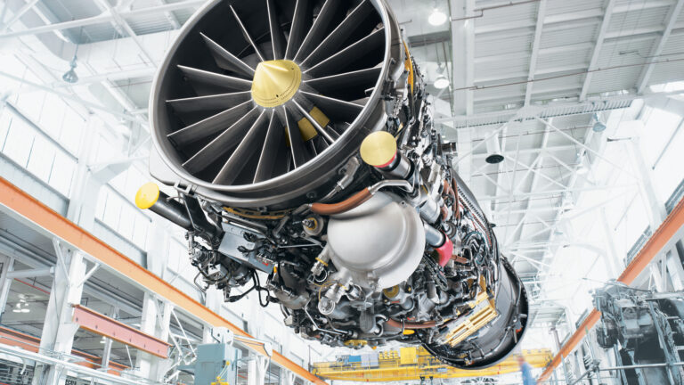 Close-up view of a large jet engine turbine fan and components inside a bright aerospace manufacturing facility, with overhead crane structure in the background.