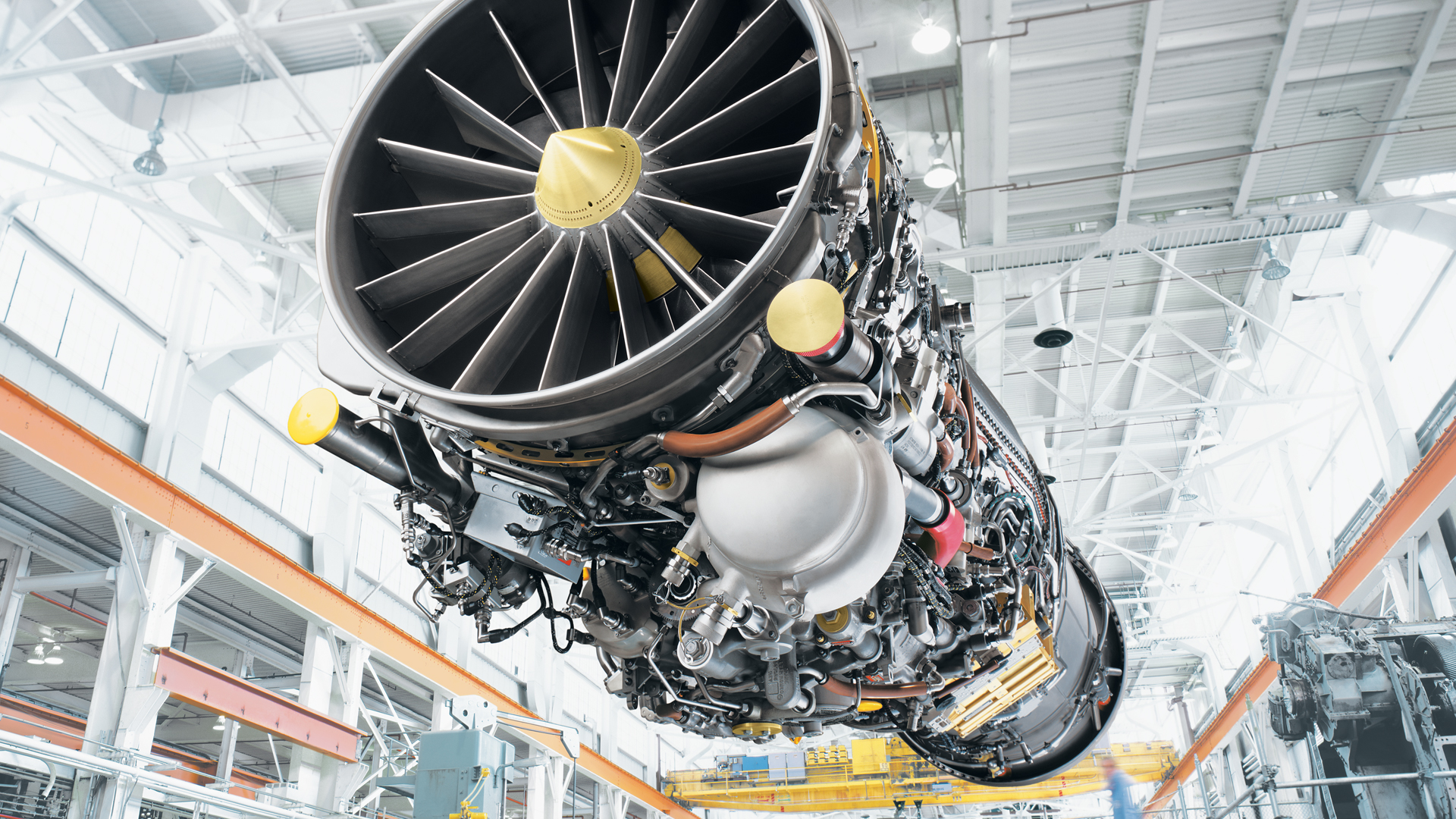 Close-up view of a large jet engine turbine fan and components inside a bright aerospace manufacturing facility, with overhead crane structure in the background.