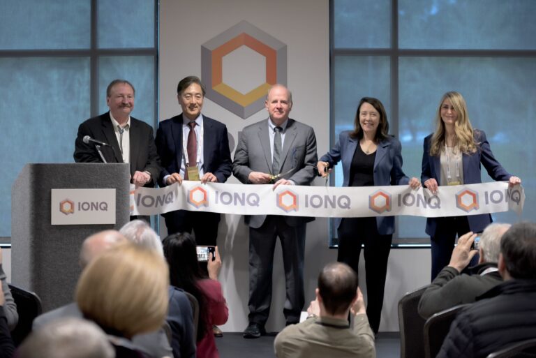 IonQ executives and partners cutting a ceremonial ribbon at the opening of a quantum computing manufacturing facility, with IonQ branding visible in the background.
