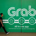 A pedestrian walks past a large green Grab advertisement billboard promoting GrabTaxi, GrabCar, and GrabHitch services.