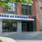 Bank of America branch entrance with blue 'Bank of America' lettering and flag logo above glass storefront windows on a brick and stone building
