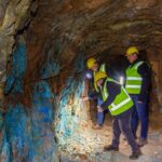 Three people in hard hats and hi-vis vests inspect blue-stained rock on the wall of an underground mine tunnel.