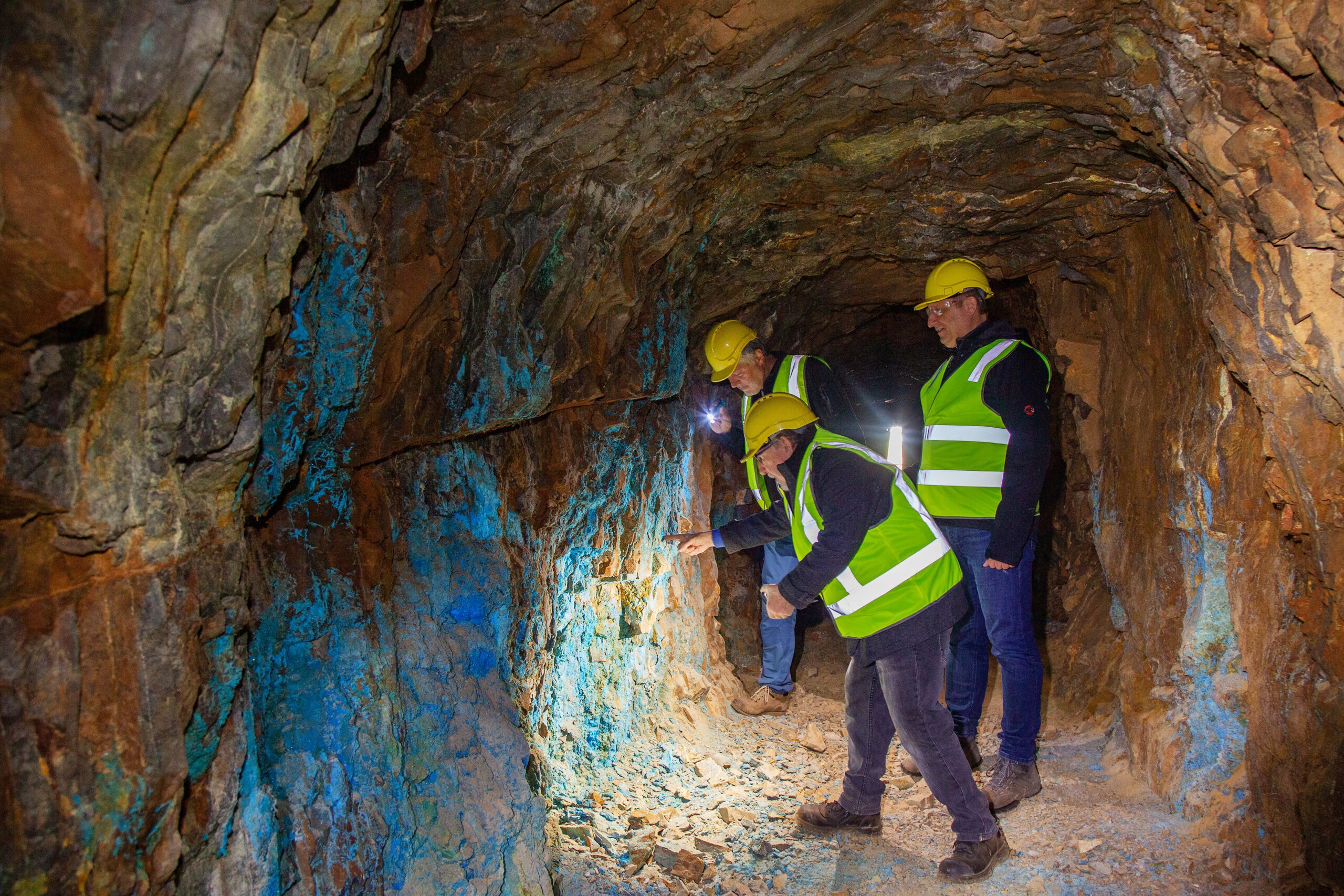 Three people in hard hats and hi-vis vests inspect blue-stained rock on the wall of an underground mine tunnel.