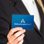 Business professional in a dark blazer holding up a blue card bearing the AdvancedAdvT logo