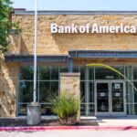 Exterior of a Bank of America branch with a large illuminated logo sign on a stone façade and an ATM beside the entrance