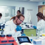Laboratory scientist in a white coat, safety glasses and blue gloves pipetting a sample while colleagues work at benches behind him