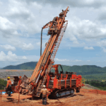 Orange drill rig with its mast raised on a red-earth mine site, with two workers in hard hats beside it and green hills behind