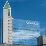 Glass office building with the Celestica name on its facade beside a stone tower with a green pointed roof, under a blue sky
