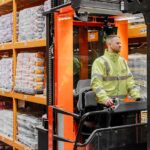 Warehouse operative in a hi-vis jacket driving a reach truck between tall orange racking stacked with bagged flooring products