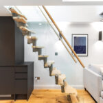 Modern open-plan apartment interior with a glass-sided floating staircase, dark kitchen units and a grey sofa