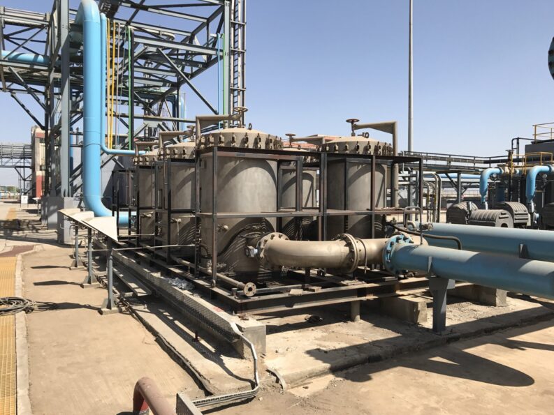 Row of large filter vessels and blue process pipes at an industrial oil and gas water treatment facility under a clear sky