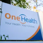 Outdoor sign for One Health reading 'Your Health. Your Choice.' in front of autumn trees and a building