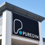 PureGym logo on a black sign mounted on the exterior of a gym building against a clear blue sky