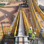 Workers in hard hats and high-visibility vests stand on a platform beside a conveyor belt carrying crushed ore at a mining processing site