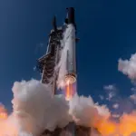 Starship rocket lifting off from its launch tower against a deep blue sky, with engines firing and large clouds of steam and flame below