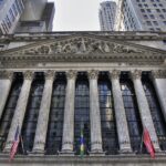 Low-angle view of the New York Stock Exchange facade with its columns, pediment sculpture and flags, surrounded by skyscrapers