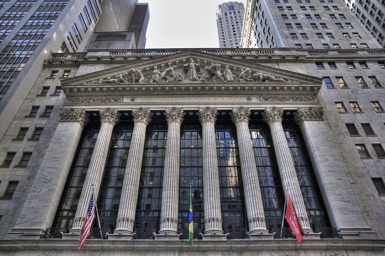 Low-angle view of the New York Stock Exchange facade with its columns, pediment sculpture and flags, surrounded by skyscrapers
