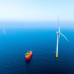 Aerial view of an offshore wind turbine and a red support vessel at sea, with another turbine and an installation vessel in the distance