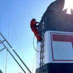 Crew member in orange overalls climbing a ship's funnel marked with a red T emblem against a blue sky
