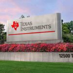 Texas Instruments sign with red TI logo above a flower bed at 12500 TI Blvd, under a pink dusk sky