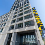 Looking up at the light stone facade of Hawthorne House, a tall student accommodation tower in Stratford, London, with a yellow and black angular screen on its side and the entrance sign at street level.