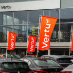 Orange Vertu flags in front of a glass-fronted Vertu dealership, with cars parked on the forecourt