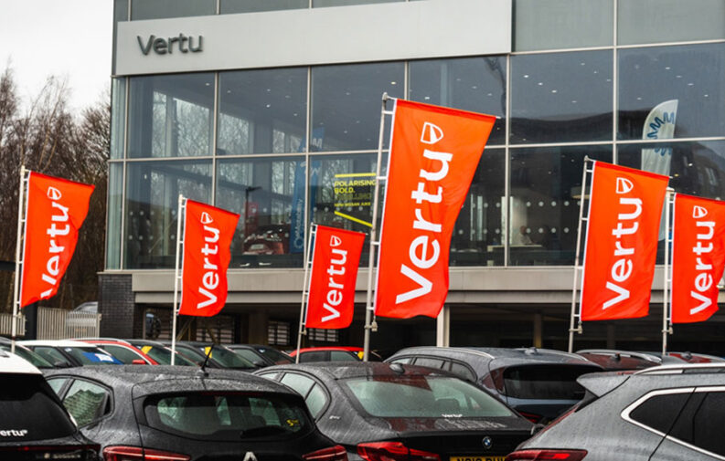 Orange Vertu flags in front of a glass-fronted Vertu dealership, with cars parked on the forecourt