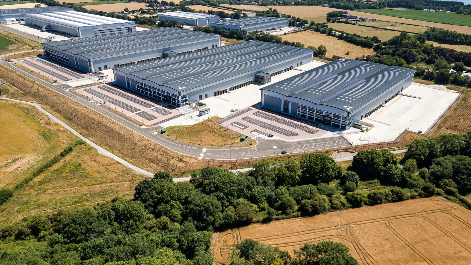 Aerial view of a modern logistics park with several large grey warehouses surrounded by farmland and trees