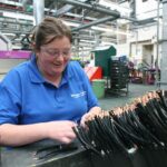 Smiling factory worker in safety glasses and a blue polo shirt assembling black electrical cables in a manufacturing facility