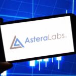 Hand holding a smartphone showing the Astera Labs logo in front of a blue stock-market candlestick chart