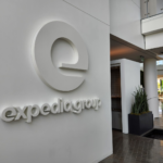 White three-dimensional Expedia Group logo and wordmark mounted on a wall in a bright office lobby