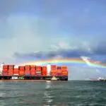 ZIM LNG-powered container ship loaded with containers passes a port with red cranes under a rainbow and stormy sky