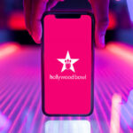 Hand holding a smartphone showing the Hollywood Bowl logo in front of a neon-lit bowling lane