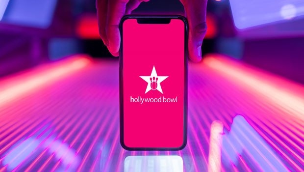 Hand holding a smartphone showing the Hollywood Bowl logo in front of a neon-lit bowling lane