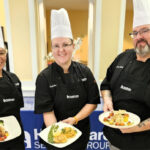 Three Healthcare Services Group chefs in black uniforms and white chef hats smile while holding plated meals.