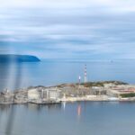 Aerial view of the Hammerfest LNG plant on Melkøya island, with storage tanks, a red-and-white mast and a small flare beside calm water.