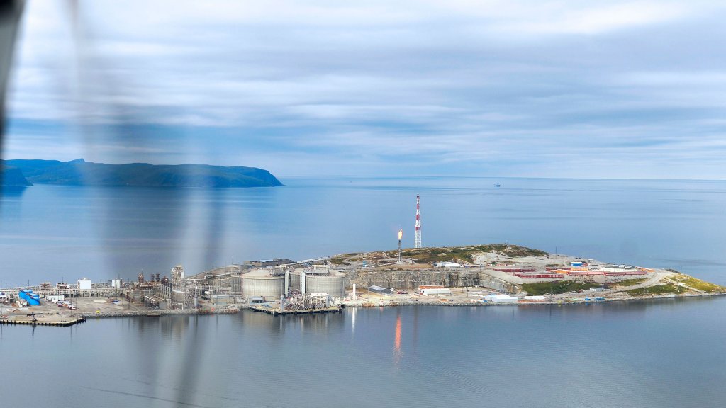 Aerial view of the Hammerfest LNG plant on Melkøya island, with storage tanks, a red-and-white mast and a small flare beside calm water.