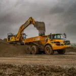 Yellow Blackwell Earthmoving excavator loading soil into a Volvo articulated dump truck on a muddy construction site under a stormy sky