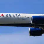 Delta Air Lines jet in flight seen from below, with the red triangle logo, DELTA lettering and blue engines against a clear sky