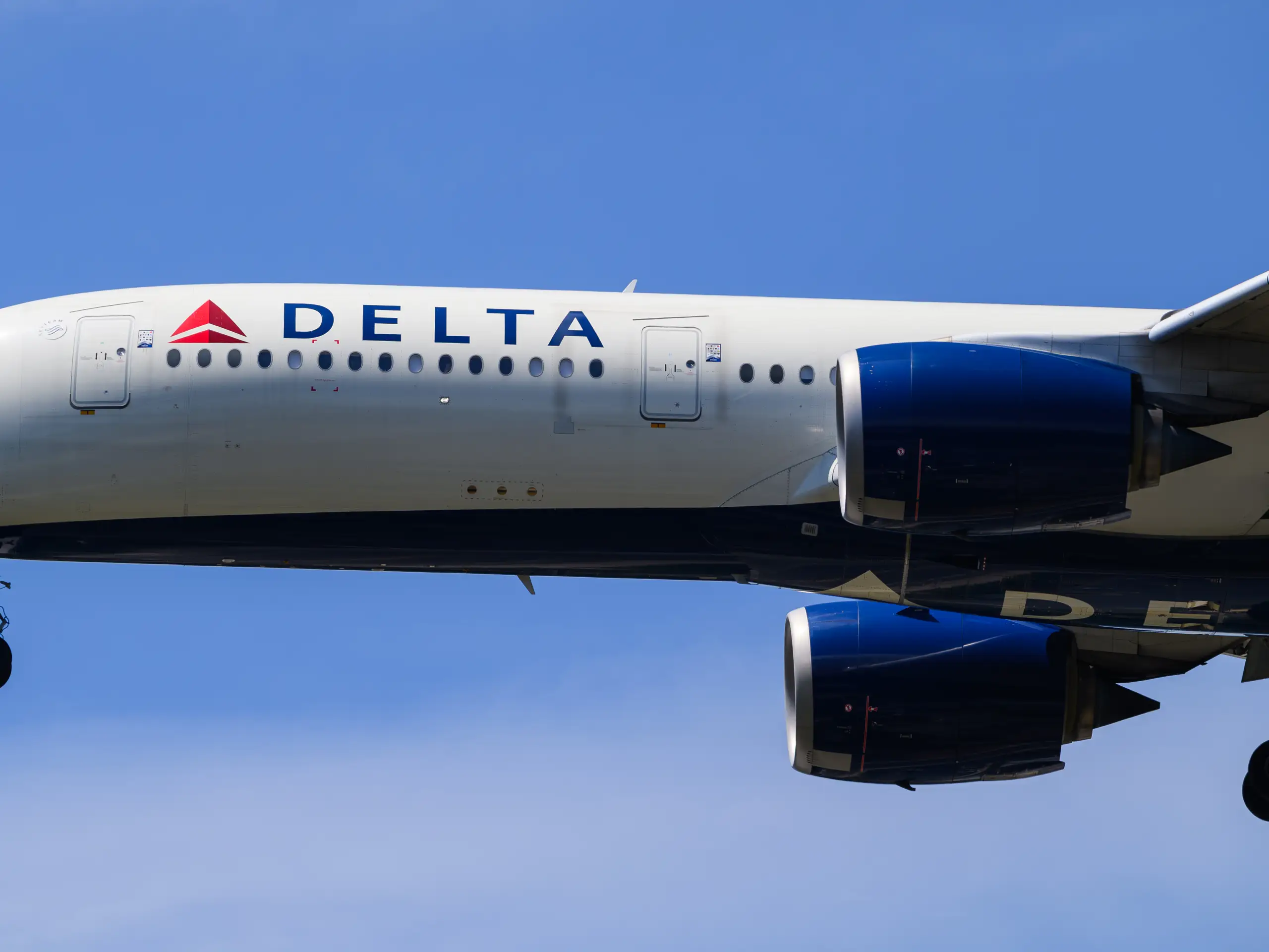 Delta Air Lines jet in flight seen from below, with the red triangle logo, DELTA lettering and blue engines against a clear sky