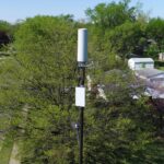 White cellular antenna and radio unit mounted on a pole above trees in a suburban neighbourhood