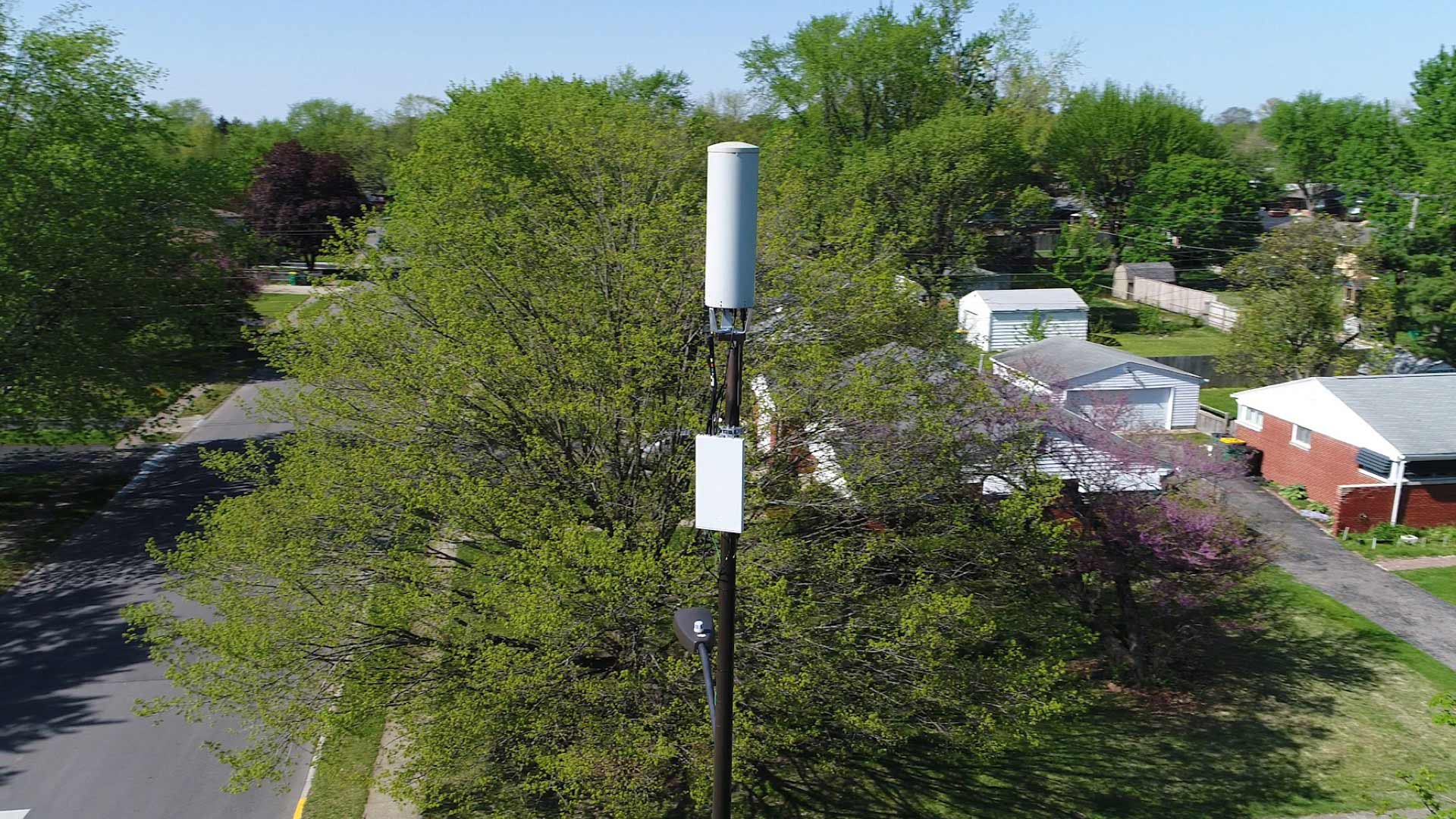 White cellular antenna and radio unit mounted on a pole above trees in a suburban neighbourhood