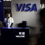 Visa staff member stands at a welcome desk beneath a large illuminated Visa logo at an exhibition booth in China, with blurred visitors passing in the foreground.