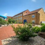 Stone and brick G&H building with a blue entrance door, seen across a brick-paved courtyard with shrubs