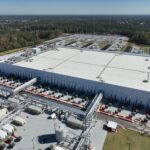 Aerial view of a large white-roofed data centre with cooling pipework, a substation and surrounding forest